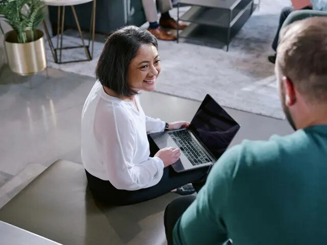 Eine Frau sitzt mit einem Laptop auf dem Schoß und spricht mit einem Mann, der neben ihr steht.